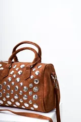 Bolso tipo bowling de gamuza color marrón con tachas plateadas. Tiene doble asa corta y correa larga desmontable.