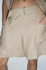 Bermuda color beige de lino, con cintura elástica, trabillas y cinturón con hebilla metálica. Presenta bolsillos de parche laterales y traseros.