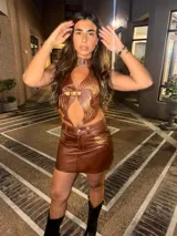 Top halter de cuero marrón con diseño de chaleco vaquero, ajustado al cuello y con cierre frontal mediante hebillas doradas. Presenta bordados en hilo dorado con motivos de llamas o plumas estilizadas en el frente.