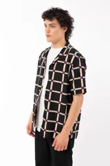 Camisa de manga corta con calce relajado y cuello tipo sastre, confeccionada en rayón. Presenta un diseño de cuadrícula beige sobre fondo negro con detalles en azul y rojo en las intersecciones.