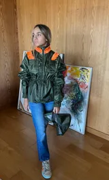 Piloto impermeable de color verde oliva con detalles de volados en color naranja sobre los hombros. Presenta capucha integrada, cintura ajustable mediante cordón con tranca, mangas voluminosas con puños elastizados y bolsillos frontales con solapa.