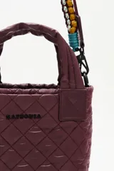 Cartera tipo tote mini acolchada, color negro brillante, con asas cortas y correa larga ajustable. La correa larga es intercambiable y presenta un diseño trenzado multicolor con tonos turquesa, amarillo y negro.