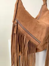 Bolso tipo hobo color tostado, hecho de cuero con flecos largos en el frente y dos bolsillos con cierre. Tiene una manija regulable con hebillas metálicas.