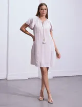 Vestido corto de lino a rayas verticales color lila y blanco, con cuello fruncido, escote en V y lazo de ajuste. Mangas cortas con puntilla de crochet.