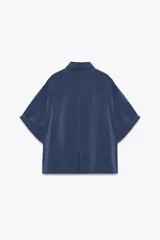 Camisa azul de cuello solapa y manga corta con dobladillo. Tiene dos bolsillos de parche en el pecho, bajo asimétrico con aberturas laterales y cierre frontal con botones ocultos.