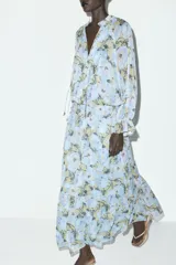 Vestido largo con estampado floral en tonos celeste, amarillo y marrón. Tiene cuello redondo con volantes y lazada, escote pico, manga larga con puño elástico con volantes, forro interior y cierre frontal con botones.