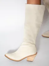 Bota alta color beige de cuero gamuzado, con punta fina y cierre lateral. Tiene un taco de madera con forma geométrica.