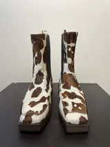Botas de cuero con estampado de vaca en tonos marrón y blanco, con elástico lateral y suela marrón.