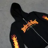 Canguro negro con cierre frontal, capucha ajustable con cordones y bolsillos tipo canguro. Estampado en el frente con el logo de la marca "Rocker Studios" en llamas y estampado de llamas con calaveras en las mangas.
