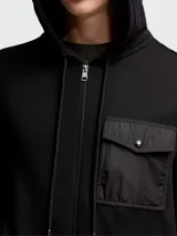 Buzo negro con capucha y cierre frontal completo. Presenta un bolsillo tipo parche en el pecho confeccionado en tejido técnico.