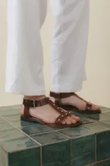 Sandalia de cuero color marrón con tiras finas, detalles de tachas metálicas y hebilla lateral.