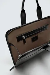 Maletín negro con acabado liso, compartimento principal con cierre de cremallera, bolsillo interior para dispositivos electrónicos de hasta 13 pulgadas, bolsillo interior con cierre de cremallera y dos asas de mano.