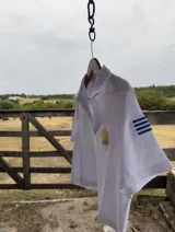 Remera tipo polo blanca de manga corta con cuello y abertura frontal. Presenta un bordado del Sol de Mayo en el pecho y la palabra "milong" bordada en azul en la manga izquierda.