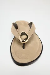 Sandalia plana color beige, con tira de serraje para el dedo y adorno metálico dorado.