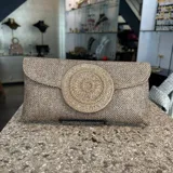 Sobre de fiesta color tostado, tejido artesanalmente en la India. Tiene aplique central circular bordado con mostacillas doradas. Incluye asa de cadena dorada para llevar al hombro.