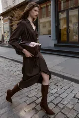 Bota alta de caña media en color marrón, confeccionada en material tipo gamuza. Presenta un diseño de punta fina, tacón alto y fino, y una tira decorativa con hebilla metálica en la parte superior de la caña. Incluye cierre lateral mediante cremallera.