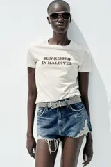 Camiseta color crema de algodón con efecto lavado, corte regular, cuello redondo, manga corta y texto marrón estampado en el frente con la frase 'Sun-Kissed in Maldives'.