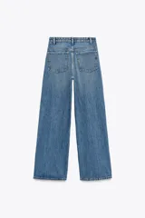 Pantalón de jean celeste de tiro alto, corte wide leg y largo hasta el tobillo. Presenta un diseño asimétrico en la cintura con cierre frontal cruzado con cremallera y botones metálicos.