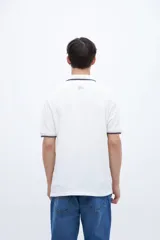 Chomba blanca de algodón con cuello clásico, media cartera de botones, bordado con la palabra 'Uniform' en el pecho y puños con rayas azul marino y rojo.
