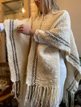 Ruana color crudo con rayas marrones tejidas en los bordes y flecos en el ruedo.