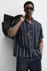 Camisa de manga corta con cuello bowling y corte relaxed fit. Diseño a rayas verticales en tonos beige y blanco. Cierre frontal con botones y bajo con aberturas laterales.