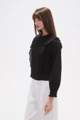 Blusa de algodón color negro con cuello a la base y detalle de volados en el frente. Posee mangas largas con puños elásticos y un panel frontal con textura fruncida.