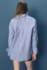 Camisa de algodón a rayas verticales azules y blancas con hilo metalizado, cuello solapa y escote pico. Manga larga acabada en puño, bajo asimétrico y cierre frontal con botones.