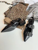 Zapato negro de cuero sintético, con punta fina, escote asimétrico y dos tiras con hebillas metálicas.