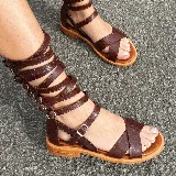 Sandalias planas de cuero color marrón, con tiras cruzadas en el empeine y múltiples tiras que se enrollan en la pierna, ajustables con hebillas metálicas.