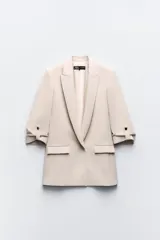 Blazer color beige de corte recto, con cuello solapa y hombreras. Tiene mangas 3/4 con trabilla y botón dorado para sujetar el dobladillo. Cuenta con bolsillos delanteros con solapa.