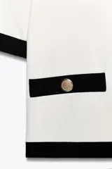 Blazer blanco de tejido de punto con cuello redondo, manga corta, bolsillos laterales y cierre frontal con botones metálicos dorados. Presenta ribetes negros en cuello, mangas, cierre y bolsillos.