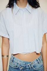 Camisa blanca corta con rayas verticales celestes, cuello solapa y mangas cortas con dobladillo. Tiene un bolsillo de parche en el pecho y cierre frontal con botones ocultos.