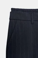 Pantalón negro con finas rayas verticales blancas, de tiro medio y corte ancho. Presenta bolsillos delanteros y traseros, cintura doble tipo bóxer con botones y cierre frontal con cremallera y ganchos metálicos. Detalle de franja marrón en los laterales.