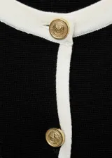 Cárdigan negro de punto fino con cuello redondo y manga corta. Presenta cierre frontal con botones dorados, ribetes blancos en contraste y dos bolsillos de ribete delanteros.