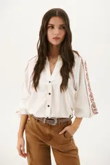 Camisa blanca de manga larga con bordados marrones en hombros y mangas, cuello en V y cierre frontal con botones.