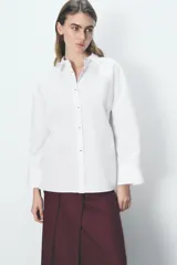 Camisa blanca oversize de popelina de algodón, con cuello solapa, manga larga con puños y cierre frontal con botones ocultos. Detalle de hilo de contraste en los botones.
