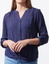 Blusa azul de gasa con cuello a la base, escote en V y abertura central con botones. Delantera con bolsillo aplicado. Largo de manga regulable con presilla y botón. Terminación opcional de nudo en ruedo.