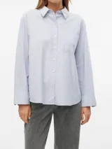 Camisa celeste de corte oversize, estilo Oxford, con cuello clásico, manga larga con puños abotonados, cierre frontal con botones y bolsillo de parche en el pecho.