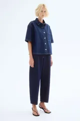 Camisa azul de denim, corte amplio, con cuello clásico, abotonadura frontal, manga corta, bolsillo de parche en el pecho y canesú en la espalda.
