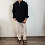 Camisa negra de manga larga con cuello mao y tapeta corta con botones de madera. El tejido presenta una textura arrugada o plisada. Las mangas están remangadas y sujetas con una presilla abotonada.