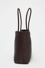 Bolso shopper alargado color marrón chocolate con diseño trenzado. Tiene doble asa de hombro y cierre superior con botón imantado. Incluye una bolsa interior con cierre fruncido.