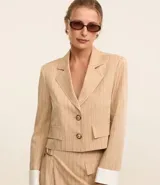 Blazer color beige con rayas verticales finas, cuello con solapa, doble botón, bolsillos delanteros y mangas largas con detalle contrastante en blanco.
