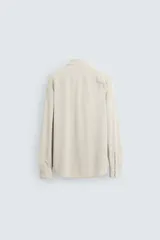 Camisa color beige de corte slim fit, confeccionada en tejido elástico con cuello italiano abierto, manga larga con puños abotonados y cierre frontal con botones.