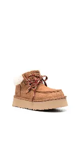Botines UGG color marrón claro, con interior de piel sintética color crema, cordones de tela con diseño geométrico en marrón, rojo y blanco, y suela de goma color beige.