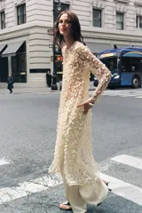Vestido largo color crema con lentejuelas, de cuello redondo y manga larga. Tiene aberturas laterales en el bajo y cierre con abertura y botón en la espalda.
