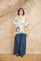 Camisa de manga corta con cuello clásico y cierre frontal con botones. Presenta un estampado con motivos florales, frutales y paisajes.