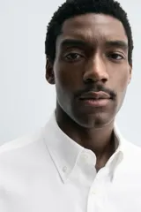 Camisa blanca de algodón, manga larga, con cuello abotonado (button-down) y cierre frontal de botones.