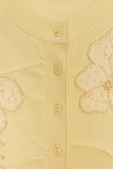 Cárdigan amarillo pálido de punto con cuello redondo, manga larga y cierre frontal con botones. Decorado con apliques florales de abalorios.