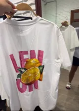 Remera blanca de corte holgado con cuello redondo y manga corta. Presenta un pequeño estampado frontal con texto en rosado y un limón cortado.