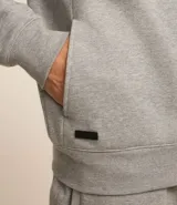 Saco deportivo gris de algodón y poliéster, con capucha ajustable con cordón, cierre frontal con cremallera, mangas largas con puños ajustados y bolsillos laterales.